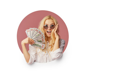 Portrait of a young shocked woman with a bundle of banknotes and text Sale. Peeping through the white hole in the background. Funny face with open mouth. Wow concept