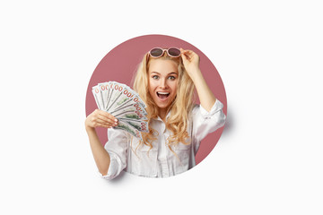 Portrait of a young shocked woman with a bundle of banknotes and text Sale. Peeping through the white hole in the background. Funny face with open mouth. Wow concept