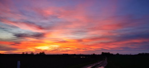 A fantastic sunset with red yellow orange and purple tones with dramatic cloud formation over green meadows with bare trees in January on the island of R&uuml;gen