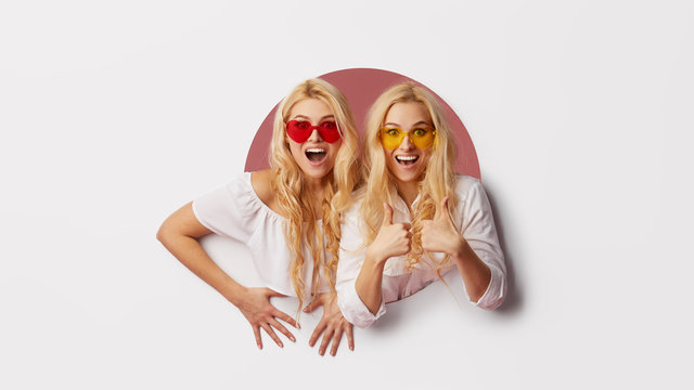Portrait of two young happy, shocked women in the heart shape sunglasses looking throth the white hole in the wall. Big sale. Funny faces. Empty space for text