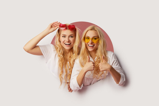 Portrait of two young happy, shocked women in the heart shape sunglasses looking throth the white hole in the wall. Big sale. Funny faces. Empty space for text