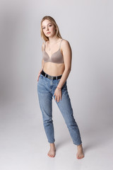 beauty portrait of sexy caucasian woman with long blond hair posing in beige lingerie and blue jeans on white background. model tests of pretty girl in bra. attractive female stands with bare feet