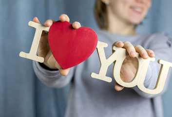 A girl in a gray sweatshirt smiles and holds in her hands a wooden inscription "I love you".