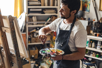 Male painter at art studio indoors micing colors looking at canvas pensive