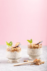Fototapeta na wymiar Yoghurt with plum, chia seeds and granola in a glass and wooden spoon on gray and pink background. side view.