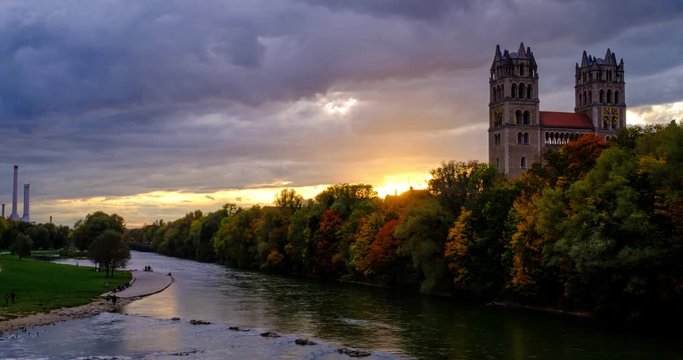 Timelapse of Munich at dramatic sunset: autumn in park of famous tourist landmark Bavarian city. View of Isar river, trees, church. Munchen, Bavaria, Germany. Camera zoom out