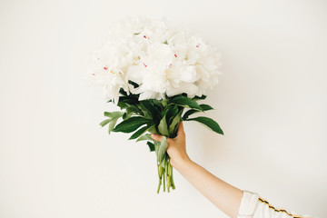 Hand holding peony bouquet on white wall background. Stylish white peonies in florist hand. Hello...