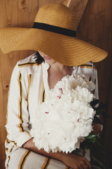 Portrait of boho woman in hat holding white peony bouquet on rustic wooden background. Stylish hipster girl in dress posing with peonies. Happy Mothers day. International Womens days