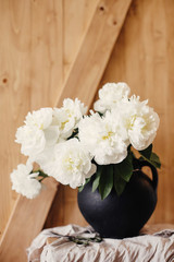 Stylish peony bouquet in black clay vase on linen fabric with scissors on rustic wooden background. White peonies rural still life. Hello spring wallpaper. Happy Mothers day. Space text