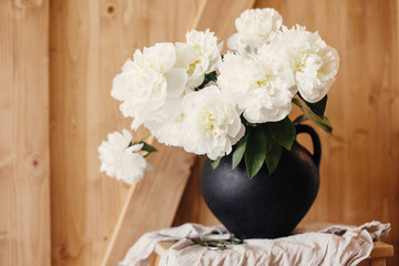 Stylish peony bouquet in black clay vase on linen fabric with scissors on rustic wooden background. White peonies rural still life. Hello spring wallpaper. Happy Mothers day. Space text