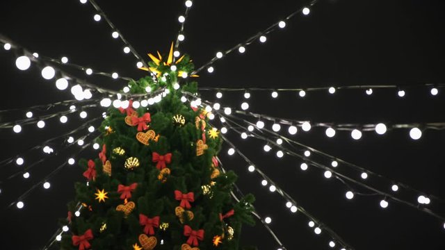 View from the bottom on the beautiful Christmas tree with colorful decorations and garland of white lights standing on the square. Concept. Beautiful snowy winter scenery of Christmas holiday