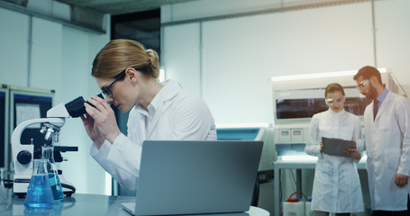 Portrait of the good looking Caucasian woman in glasses and whte robe doing some investigation at the laptop computer and analysis while looking in the microscope in the laboratory.
