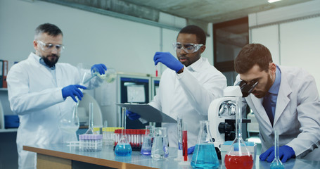 Portrait shot of the Caucasian man scientist looking in the microscope while investigating something in the laboratory, while his male mixed-races co-workers talking on the background.