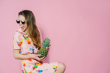 Obraz na płótnie Canvas Beautiful woman in dress with printed friuts posing with pineapple on pink background. Pineapple in lady's hands. Trendy and stylish fashion.