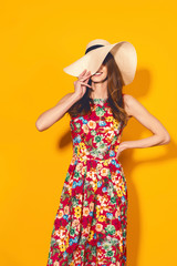 Girl in floral dress and hat close half of her face and emotionally poses on the yellow background.