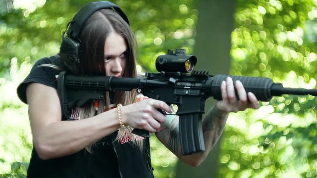 An attractive female shooting an assault rifle with red dot / reflex sight in the forest, ejecting a smoking shell from the chamber in slow motion.