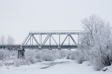 Winter landscape with railroad bridge over frozen river, snow-covered trees, hoarfrost.