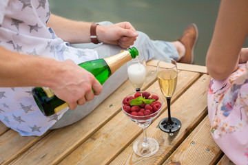 Romantic date surprise. A young guy and a girl on a wooden pier. The guy pours champagne into the glasses.
