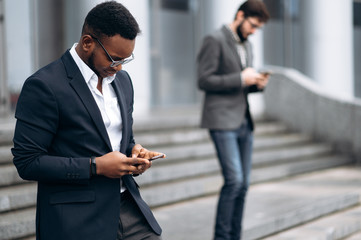 Young handsome african american businessman is walking on stairs and typing something on his phone with smile, outdoors