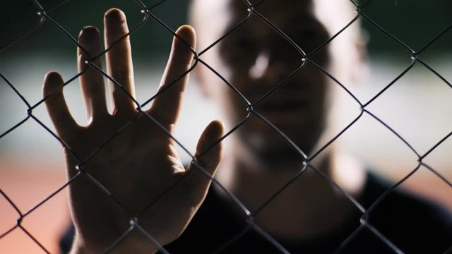 Caucasian man staying in protected place or prison holding hand on metal fence looking close-up at camera. Hopelessness concept.