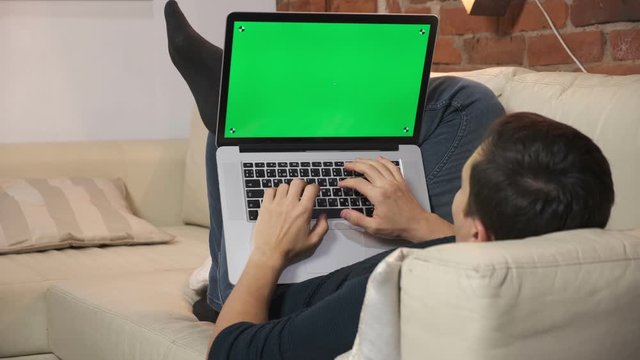 The person at the table is browsing the Internet on a green laptop screen. In a well-lit, cozy apartment. A man works in a loft office