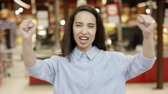 Pretty girl in casual shirt showing 'yes' winner gesture celebrating victory, smiling joyfully in excitement, in supermarket with people moving in blurred background.