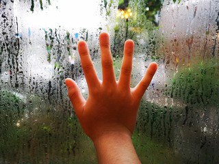 child's hand is placed on the window glass and there are water drops on the outside.
