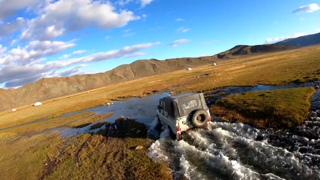 Aerial FPV, drone shot, around a 4x4 offroad truck, crossing a stream in the Gobi desert, in the Altai mountains, sunny day, in Mongolia