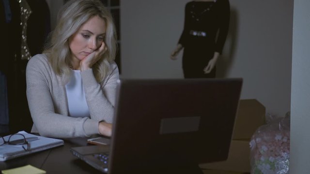 Small business owner working from home office late at night. Tired young woman sitting at desk looking at laptop computer