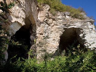 Two large cave openings on limestone bluff along the Mississippi River in the Alton and Grafton Illinois area with overgrown weeds and trees partially covering entrances on a sunny day with blue skies