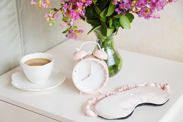 Alarm clock closeup, a cup of coffee and bouquet of pink flowers background in the morning sunlight