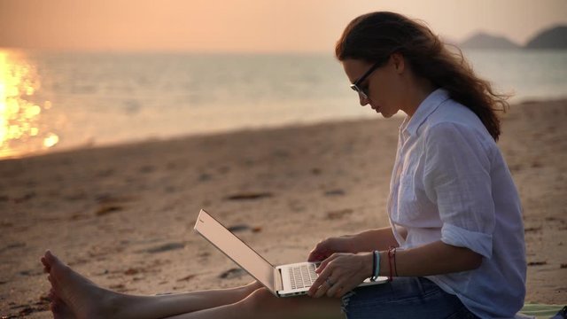A young woman writer sitting on the beach at the sunset time with her laptop and typing a text.