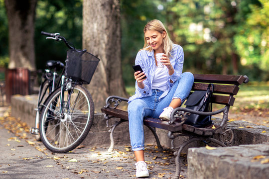 Blonde young woman sitting on the bench looking on mobile phone and drinking coffee to go in park with bicycle on the background - Powered by Adobe