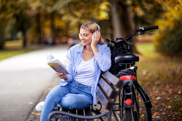 Fototapeta na wymiar Blonde girl listen music with headphones over her head and read book in park. She is sitting on bench with bicycle beside her