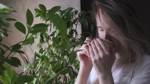 4A young girl walks to the window near home plants and drinks coffee. Freedom concept, idea. 4K slow motion