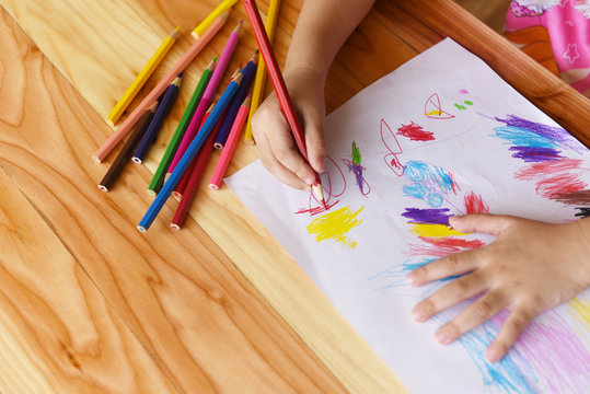 Girl painting on paper sheet with colour pencils on the wooden table at home - child kid doing drawing picture and colorful crayon