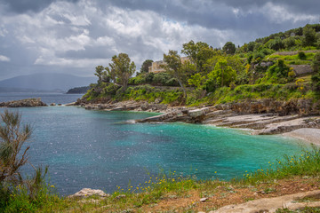 Fototapeta premium Beautiful landscape – sea lagoon with turquoise calm water, stones and rocks on the beach, cloudy sky and green trees. Corfu Island, Greece, Kassiopi village