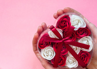 A gift in the hand. Valentine's day gift on a pink background: a lot of red and white roses in a heart-shaped package, tied with a red ribbon. The concept of love/ copy space