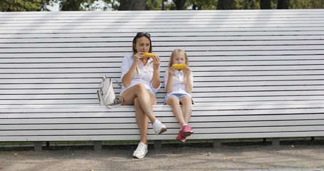 Young mother with her daughter eating boiled corn in the park in summer. An attractive woman and girl are relaxing outdoors.