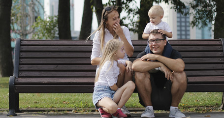 Happy family resting together in the park. Parents with children sit on the bench and wave their hands.