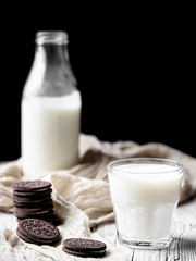 A glass with milk and several chocolate cookies on a wooden surface. Close-up. Bottle of milk in the background. Natural dairy products. Vertical frame, space for text. Shallow depth of field.
