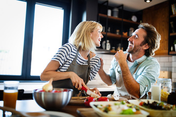 Beautiful young couple is talking and smiling while cooking healthy food in kitchen at home - obrazy, fototapety, plakaty