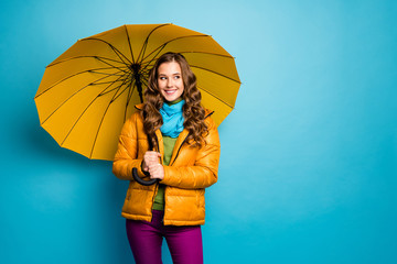 Photo of pretty lady look side empty space hold big open umbrella enjoy sunny day walk street wear yellow overcoat blue scarf violet trousers isolated blue color background