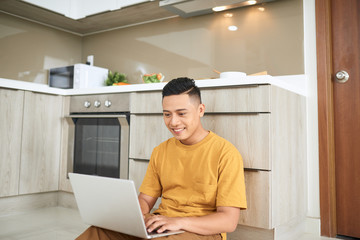 Fototapeta na wymiar attentive asian man using laptop while sitting on floor in kitchen