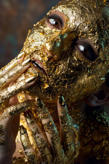 Girl with a mask on her face made of gold leaf. Gloomy studio portrait of a brunette on an abstract background.