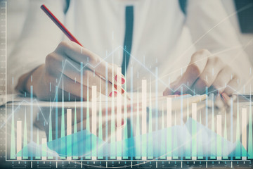 A woman hands writing information about stock market in notepad. Forex chart holograms in front. Concept of research.