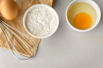 Fototapeta na wymiar Getting ready to bake, whisk, bowl of flour, small bowl of cracked egg white and egg yolk, with raw egg on grey stone background and a brown crumpled paper.