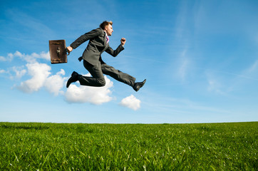 Energetic businessman jumping with briefcase outdoors across wide green meadow