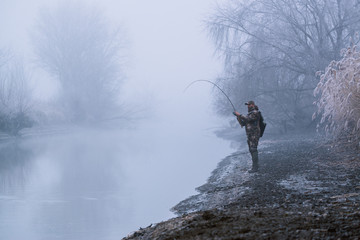 Fisher man fishing with spinning rod on a river bank at misty foggy winter, spin fishing, prey...