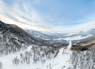 Aerial Picturesque panoramic landscape in the Altai mountains with snow-capped peaks under a blue sky with clouds in winter with gondola cableway and booths on ski resort. White snow and calm.